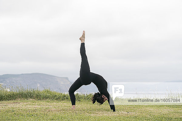 Flexible junge Frau übt Yoga auf Gras gegen den Himmel