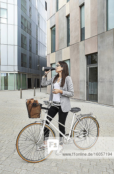 Businesswoman drinking water while standing with bicycle on city street