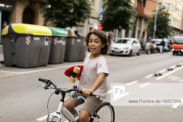 Carefree boy holding skateboard while riding bicycle on street