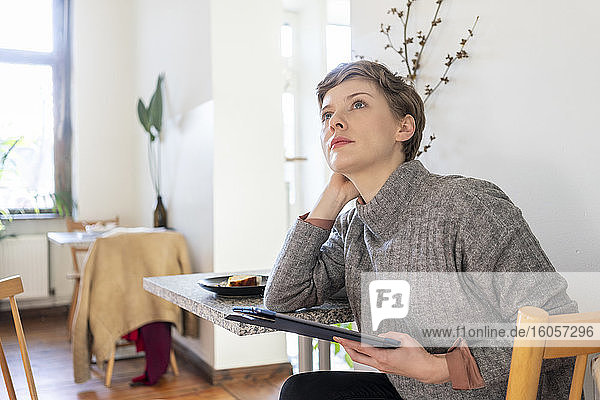 Nachdenkliche Frau  die ein digitales Tablet hält und sich auf einen Tisch in einem Café stützt