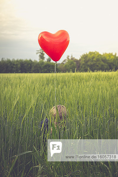 Kleiner Junge mit rotem herzförmigen Luftballon  der sich in einem Feld versteckt
