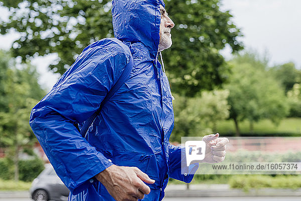 Älterer Mann in blauem Regenmantel läuft gegen Bäume im Park