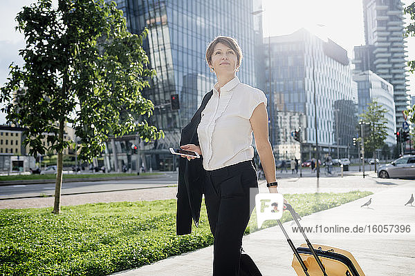 Confident businesswoman holding smart phone and suitcase while walking in city