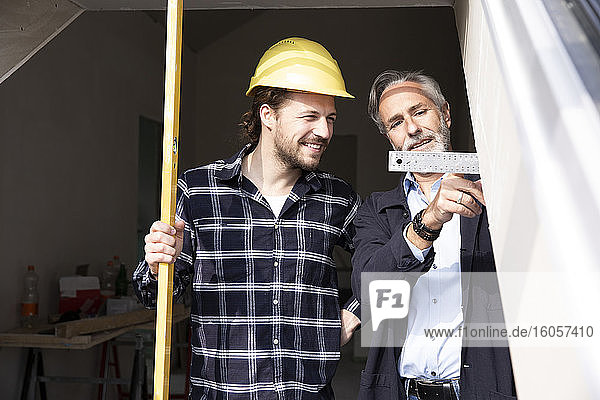 Lächelnder Bauarbeiter  der die vom Architekten beim Bau eines Hauses gezeigten Maße durch ein Fenster betrachtet