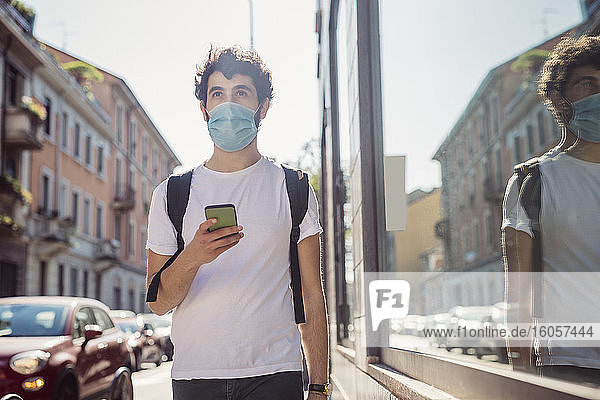 Man wearing mask holding smart phone while walking in city