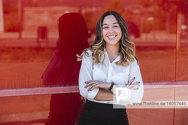 Lächelnde Geschäftsfrau mit verschränkten Armen vor einer roten Wand in der Stadt