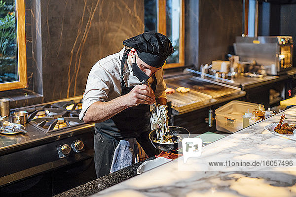 Koch mit Schutzmaske bei der Zubereitung eines Gerichts in einer Bratpfanne in einer Restaurantküche