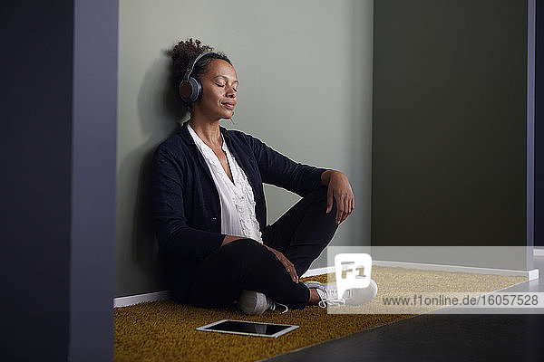 Porträt einer Geschäftsfrau  die im Büro auf dem Boden sitzt und mit Kopfhörern und digitalem Tablet Musik hört