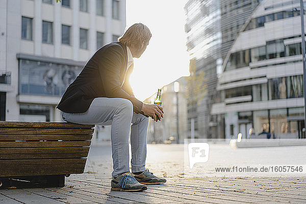 Businessman sitting on a bench in the city at sunset with a bottle of beer