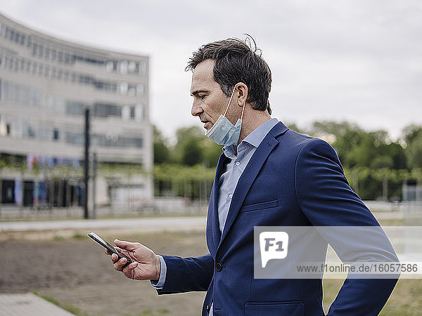 Mature businessman with protective face mask using smartphone in the city