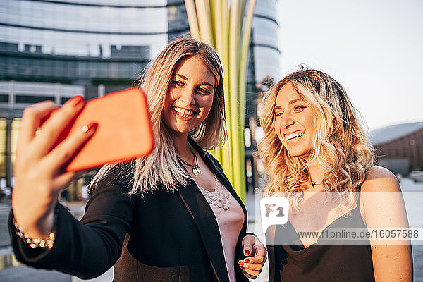 Lächelnde Geschäftsfrau  die ein Selfie mit einer Mitarbeiterin macht  während sie in der Stadt bei Sonnenuntergang steht