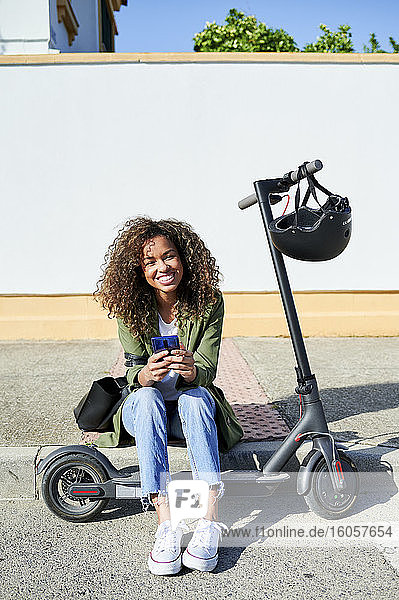 Glückliche junge Frau  die ihr Smartphone in der Hand hält  während sie auf dem Bürgersteig mit einem Elektroroller sitzt