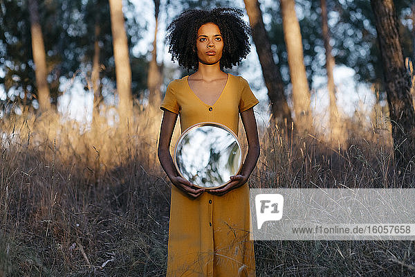 Junge Frau mit Afro-Haar  die einen Spiegel hält  während sie im Wald bei Sonnenuntergang steht