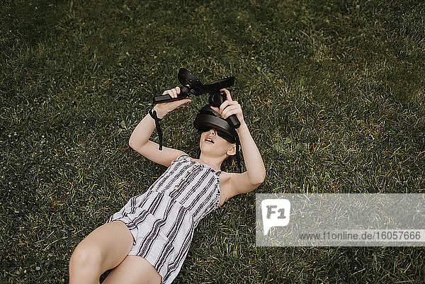 Mädchen  das einen Virtual-Reality-Simulator trägt und ein Videospiel spielt  während es auf einer Wiese im Hof liegt