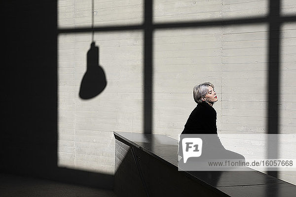 Weibliche Fachkraft im schwarzen Anzug entspannt sich auf einer Stützmauer mit Sonnenlicht und Schatten im Hintergrund im Büro