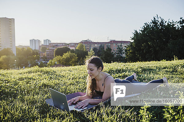 Sportliche junge Frau  die einen Laptop benutzt  während sie auf einer Übungsmatte im Stadtpark liegt