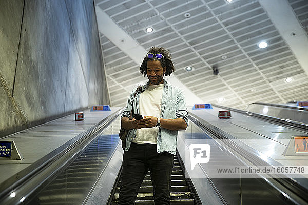 Junger trendiger Mann lächelt  während er sein Smartphone auf einer Rolltreppe in einer U-Bahn-Station benutzt