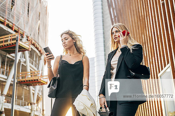 Female colleagues using smart phones while standing against buildings in city