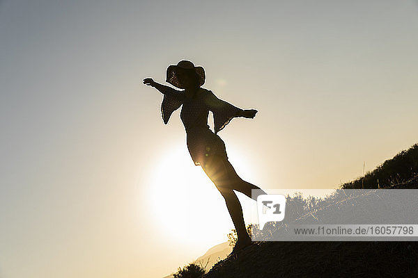 Junge Frau steht mit ausgestreckten Armen auf einem Hügel vor einem klaren Himmel