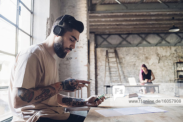 Kreativer junger Geschäftsmann mit Kopfhörern und Smartphone im Loftbüro