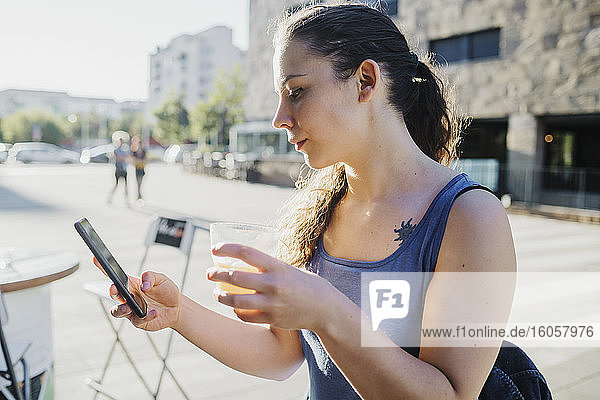 Sportliche junge Frau  die ihr Smartphone benutzt und ein Getränk in einem Straßencafé hält