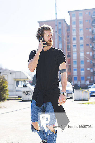 Gutaussehender Mann  der über sein Smartphone spricht  während er an einem sonnigen Tag auf der Straße geht