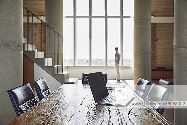 Laptop on wooden table in a loft flat with man at the window in background