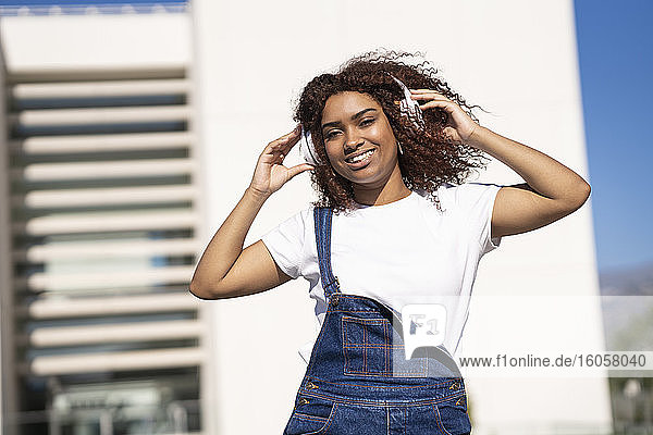 Lächelnde Afro-Frau  die Musik über Kopfhörer hört  während sie an einem Gebäude steht