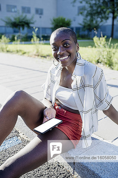 Lächelnde junge Frau mit rasiertem Kopf  die ein Mobiltelefon benutzt  während sie auf einem Fußweg in der Stadt sitzt