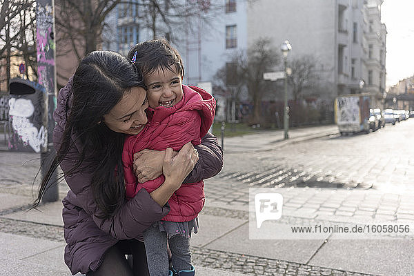Glückliche Frau  die ihre Tochter umarmt  während sie auf einem Fußweg in der Stadt kniet