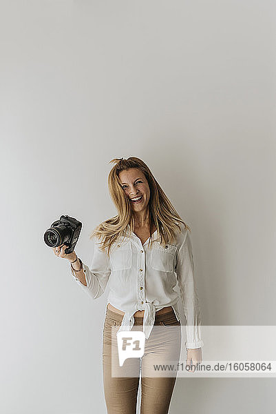 Glückliche Frau  die eine Kamera hält  während sie gegen eine weiße Wand zu Hause steht