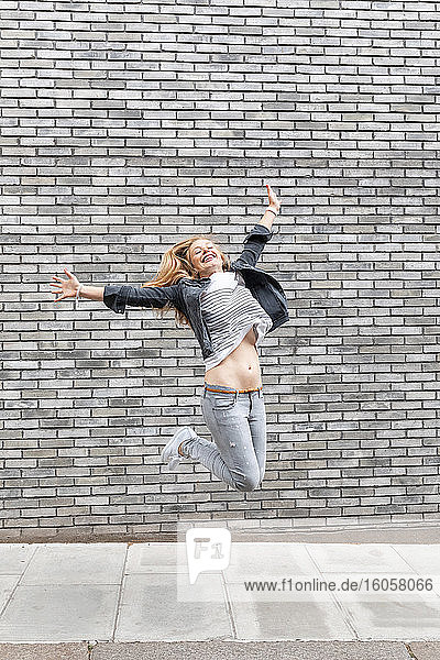 Happy woman with arms outstretched jumping on footpath against gray brick wall