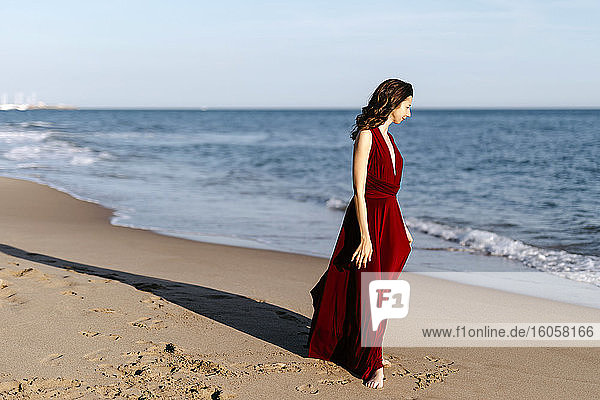 Zarte Frau in rotem Kleid geht am Meer spazieren und genießt die Sonne