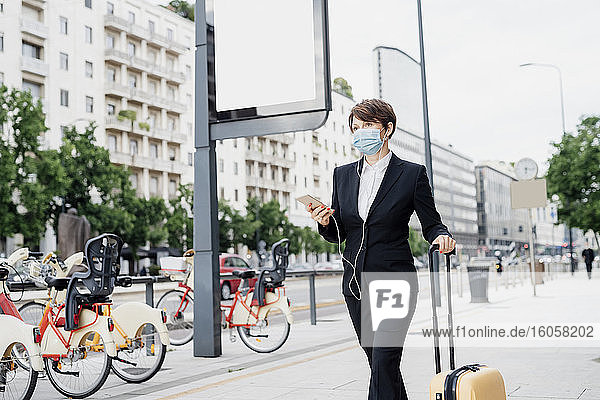 Businesswoman wearing mask listening music while walking with suitcase on sidewalk in city