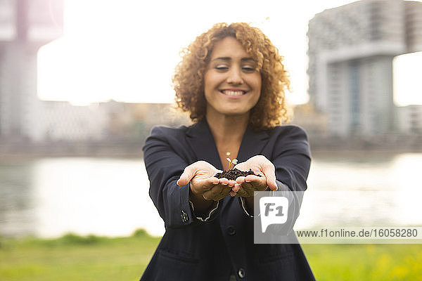 Lächelnde Geschäftsfrau hält einen Bäumchen  während sie am Fluss in der Stadt steht