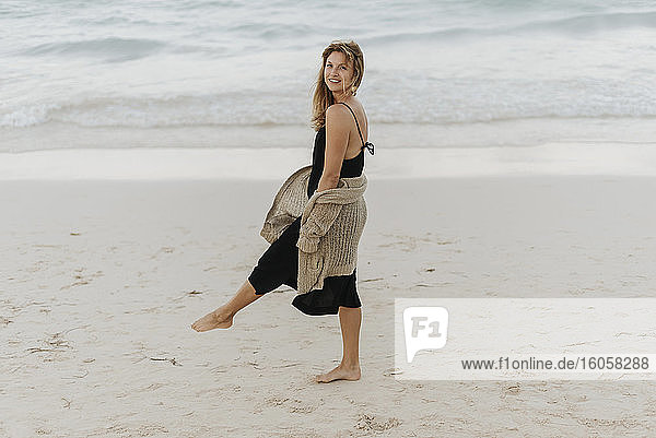Lächelnde erwachsene Frau in schwarzem Kleid  die am Strand spazieren geht