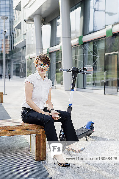 Smiling businesswoman with electric push scooter sitting on seat in city