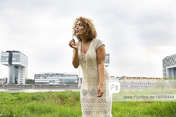 Mid erwachsene Frau mit lockigem Haar bläst Blume  während stehend auf grasbewachsenen Land gegen Himmel