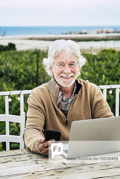 Smiling senior man holding smart phone while sitting with laptop at table