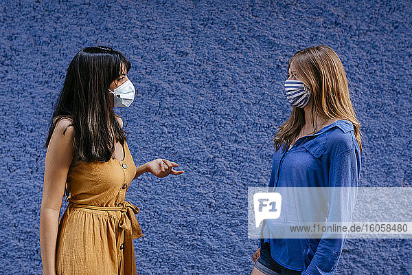 Maskierte Freundinnen unterhalten sich auf Abstand an einer blauen Wand in der Stadt