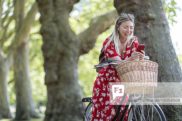 Cheerful woman using smart phone while leaning on bicycle at public park