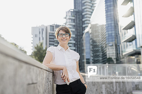 Lächelnde Geschäftsfrau mit Brille an einer Stützmauer vor einem Gebäude in der Stadt