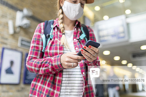 Nahaufnahme einer Frau mit Maske  die ein Smartphone benutzt  während sie an einer U-Bahn-Station steht