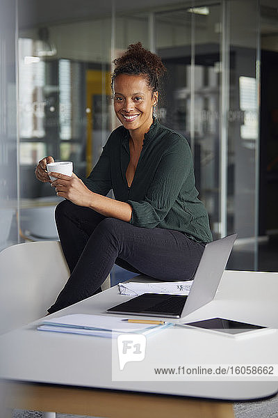 Porträt einer lächelnden Geschäftsfrau  die mit einer Tasse Kaffee am Schreibtisch sitzt und eine Pause macht