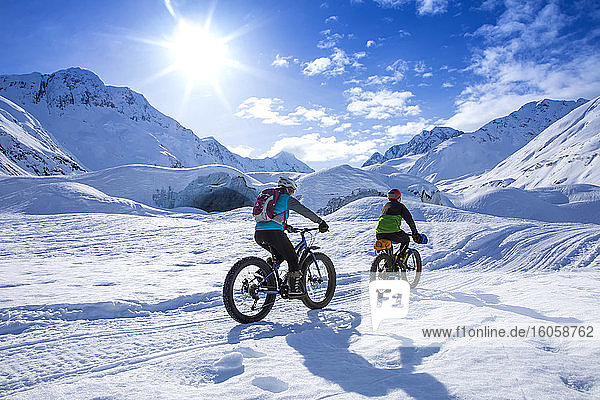 Zwei fette Frauen radeln an einem sonnigen Wintertag vor dem Skookum-Gletscher  Chugach National Forest  Alaska  und pumpen mit der Faust  während sie vorbeifährt; Süd-Zentral-Alaska; Alaska  Vereinigte Staaten von Amerika