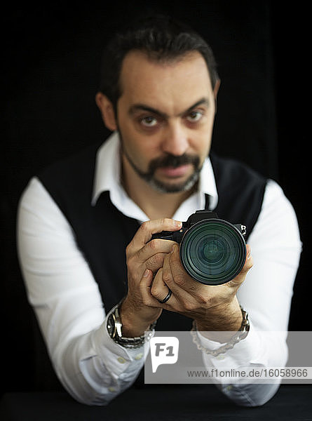 Portrait of a male photographer standing with his camera against a black background  focus on the camera in the foreground; Studio