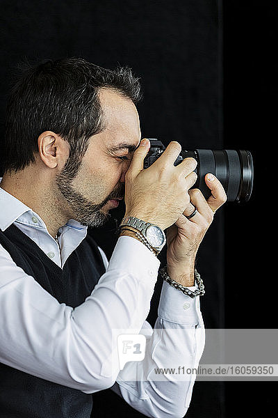 Porträt eines männlichen Fotografen  der vor einem schwarzen Hintergrund steht und seine Kamera benutzt; Studio