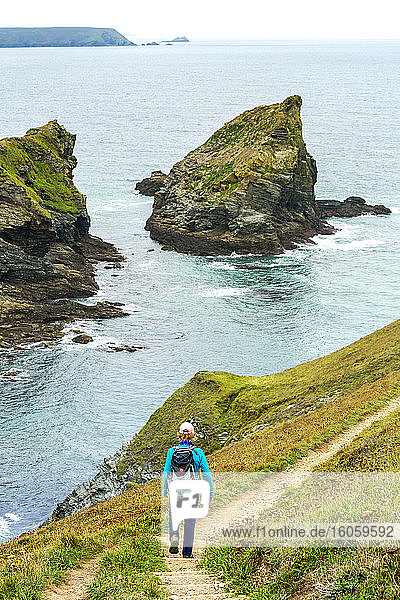 Female hiker walking down a stone stair trail with rocky formations along a cliff shoreline; Cornwall County  England