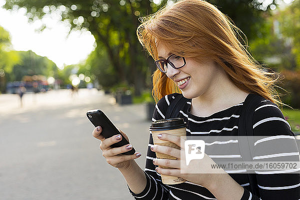 Eine junge Studentin steht mit einer Kaffeetasse in der Hand und benutzt ihr Smartphone auf dem Campus; Edmonton  Alberta  Kanada
