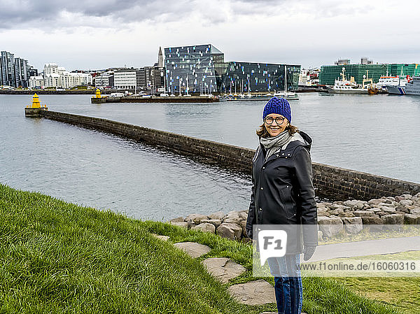 A woman stands at the waterfront with the Harpa Concert Hall and Conference Center in the background; Rejkjavik  Reykjavik  Iceland